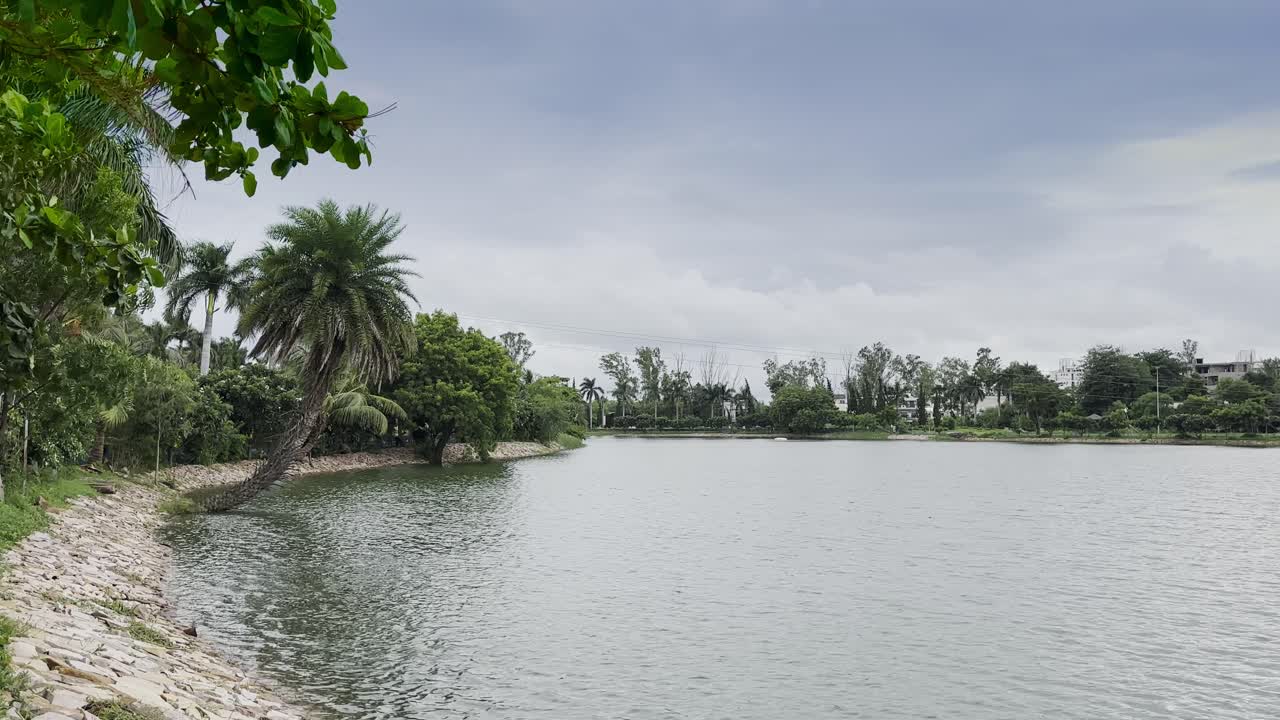 Immagine statica di un ampio lago calmo circondato da palme e vegetazione fitta. La costa si curva naturalmente con argini in pietra, creando un paesaggio tropicale tranquillo sotto un cielo nuvoloso