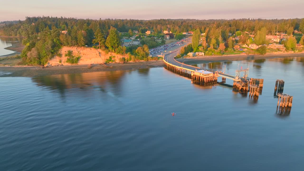 vista aérea de drones dando vueltas a la izquierda de dos personas en kayak de mar remando en una bahía cerca de seattle, washington al amanecer, revelando la ciudad de la comunidad de southworth y el muelle de la terminal de ferry