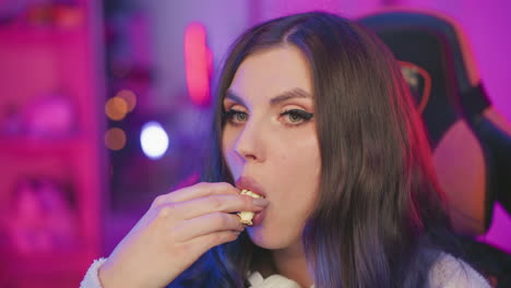 portrait view of female artist sitting in gaming chair eating popcorn with eyes closed, adjusting hair slightly under moody purple lighting, relaxed indoor vibe with glowing neon background ambiance