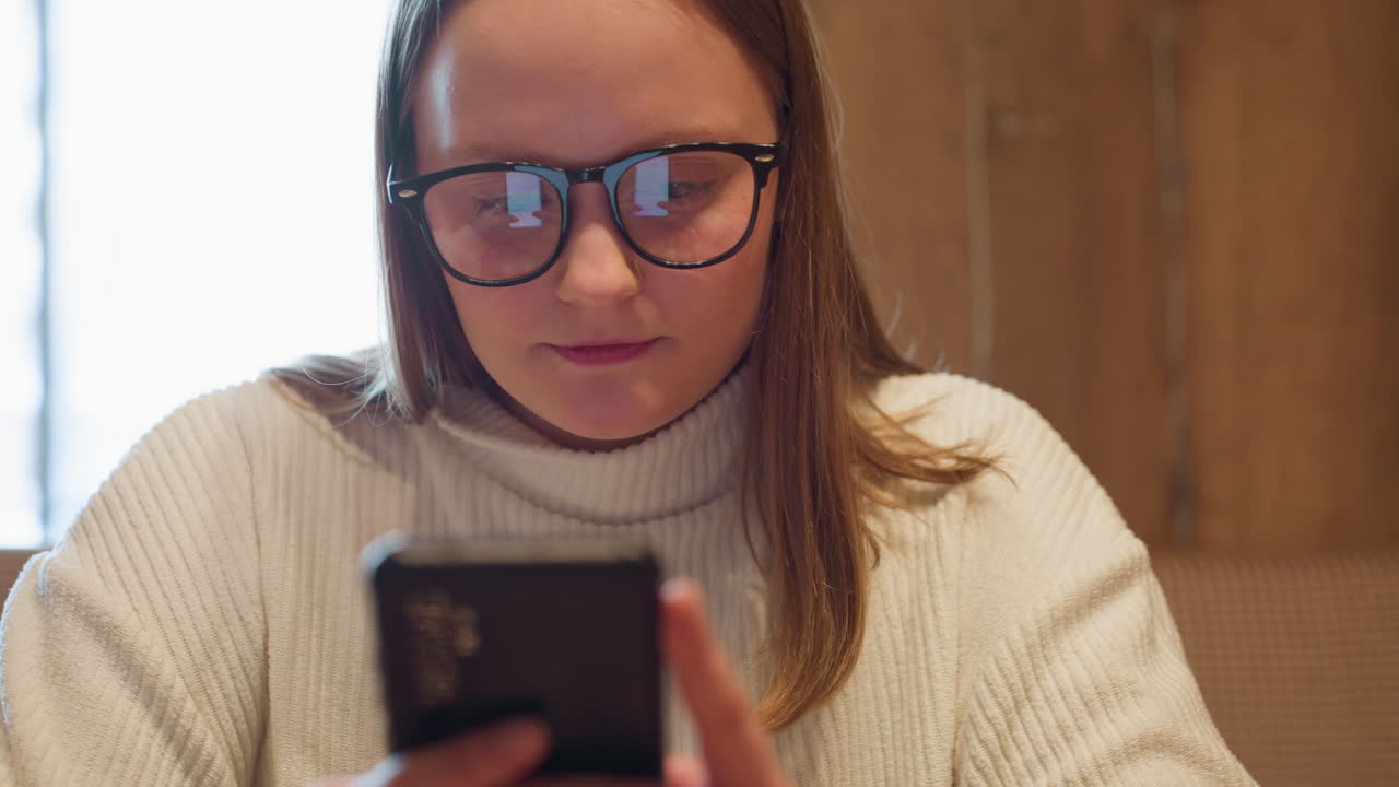 Close up of young lady in white ribbed sweater and glasses looking at smartphone while chatting, with phone screen reflected in lenses, seated in cozy indoor setting with soft lighting
