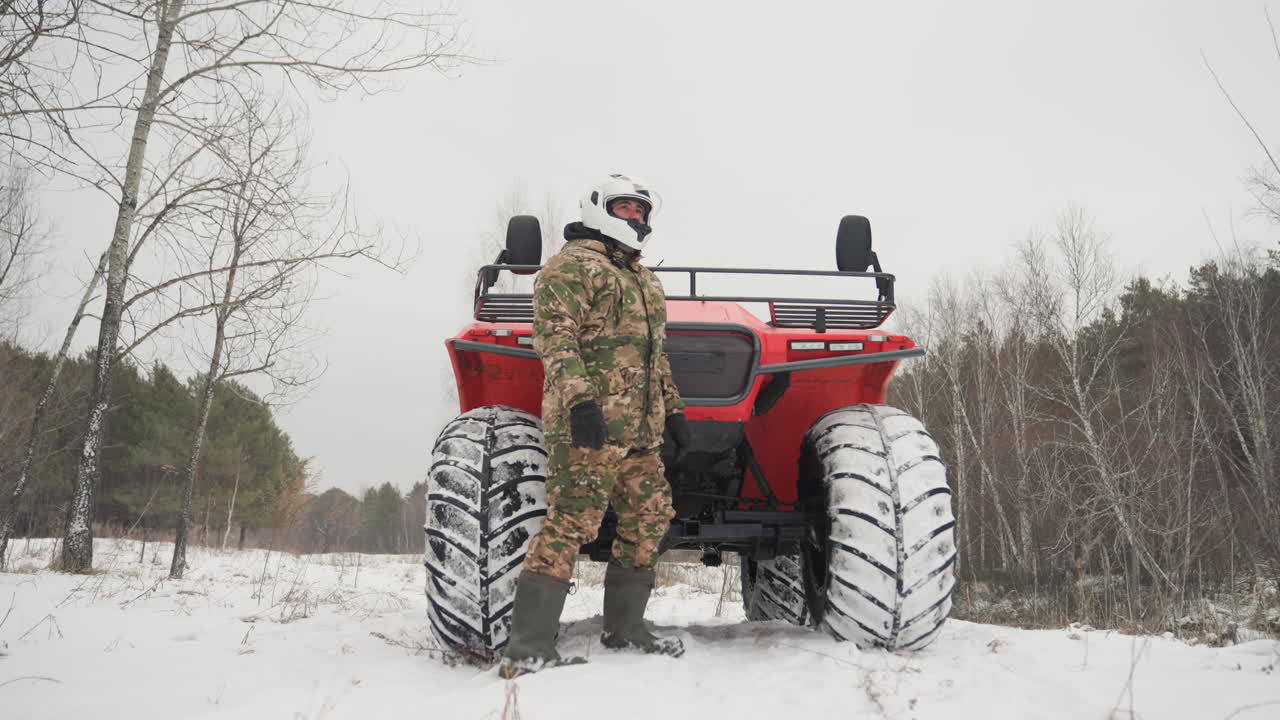 Uomo in mimetica accanto a un ATV rosso su un sentiero innevato, pilota con casco si ferma e ispeziona il veicolo ai margini di un bosco di betulle e pini, pneumatici robusti e attrezzatura invernale visibili suggeriscono caccia e manutenzione fuoristrada.