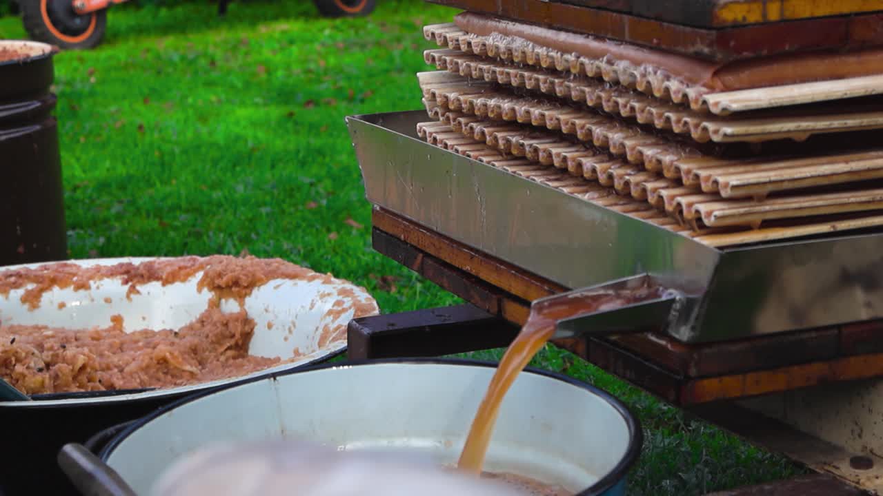 Golden brown apple juice flowing from a home garden press into a bucket in slow motion and a male hand takes the bucket away. Shallow depth of field and bokeh blurry green grass in the background