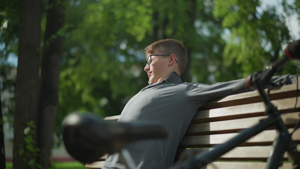 un niño con gafas extiende los brazos mientras está sentado en un banco de madera, la bicicleta estacionada es visible a su lado, con árboles y vegetación exuberante en el fondo borroso