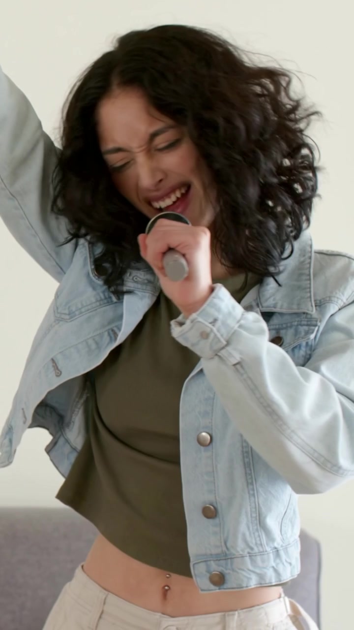 Video verticale: Donna con microfono che esegue un assolo vocale a casa, facendo pratica, indossando una giacca di jeans.