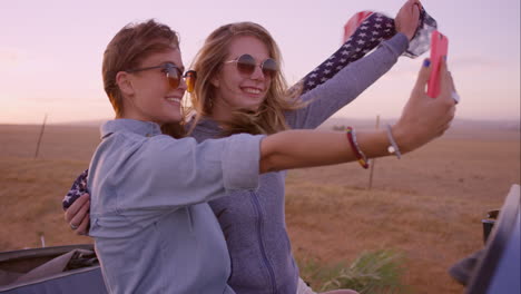 beautiful girl friends taking selfies on road trip at sunset with vintage car