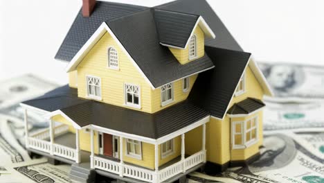 yellow model house sitting on a bed of one hundred dollar bills isolated on a white background represents real estate investment, home ownership, and the financial aspects of buying a property