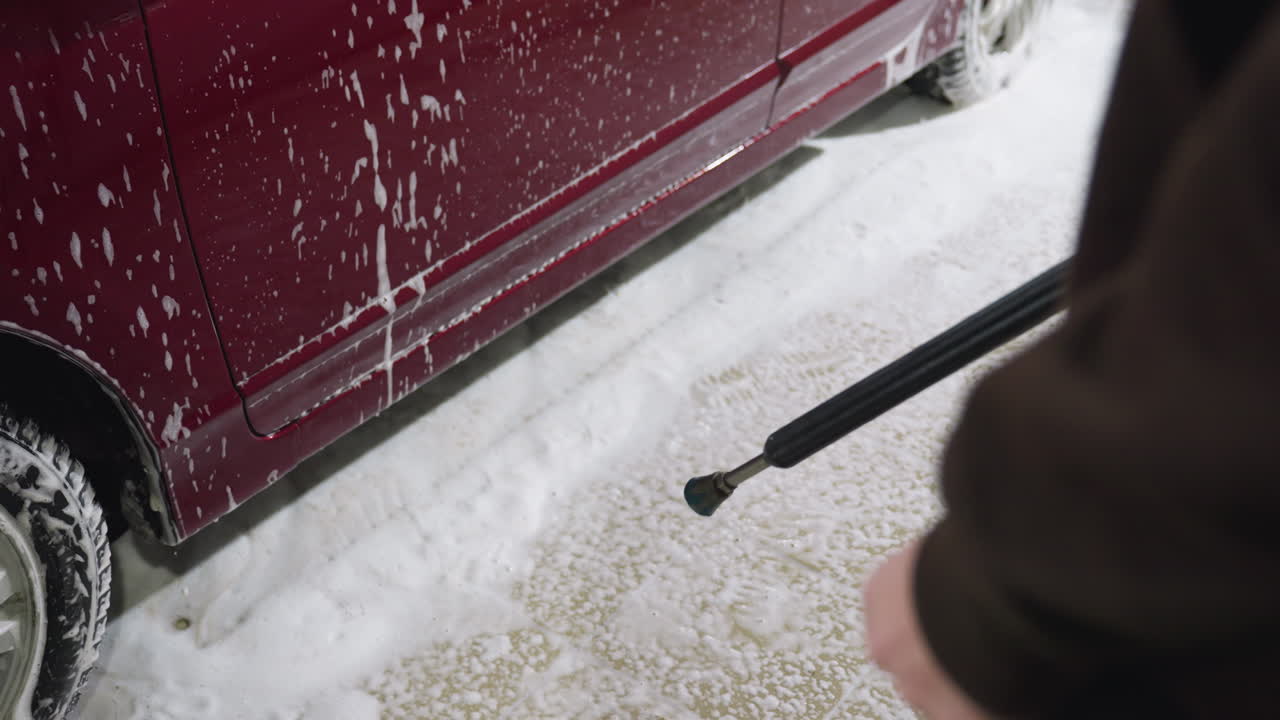 Close up of person holding spray hose directed at red car side door during washing process with foam and water flowing down vehicle exterior inside indoor garage environment during cleaning action