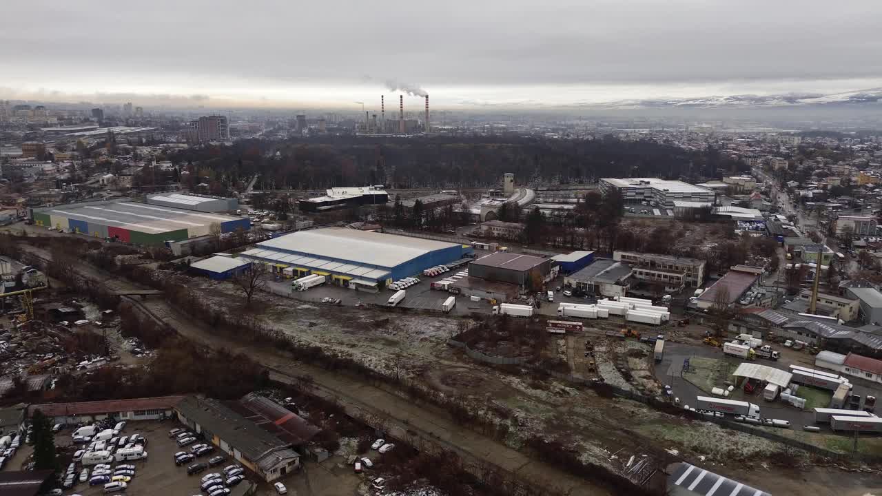 Drone footage over the industrial district of Sofia, Bulgaria, featuring warehouses, freight trucks, parked cars, and three towering chimneys emitting smoke under a dreary, overcast sky.