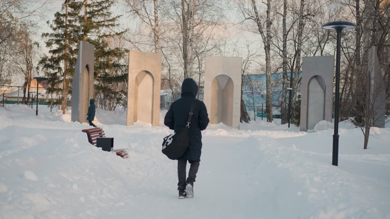 Rear view of person wearing hoodie and carrying bag walking through snowy park path, with another person crossing behind monumental arch structures and leafless trees under soft winter light