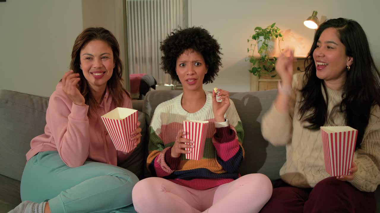 Forskellige veninder sidder i sofaen derhjemme og spiser popcorn fra spande, peger på skærmen og griner