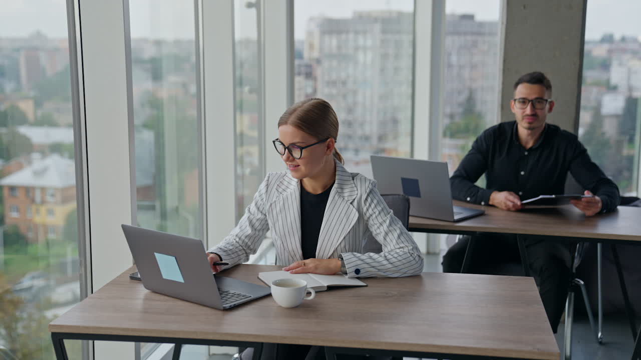 People busy with their work in the office sitting at desks. Employees solving work issues in big light office with panoramic windows with blurred cityscape.