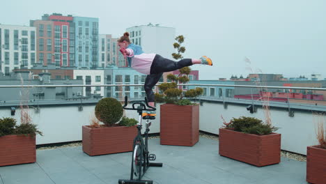 athletic girl performing aerobic riding training exercises on cycling stationary bike on house roof