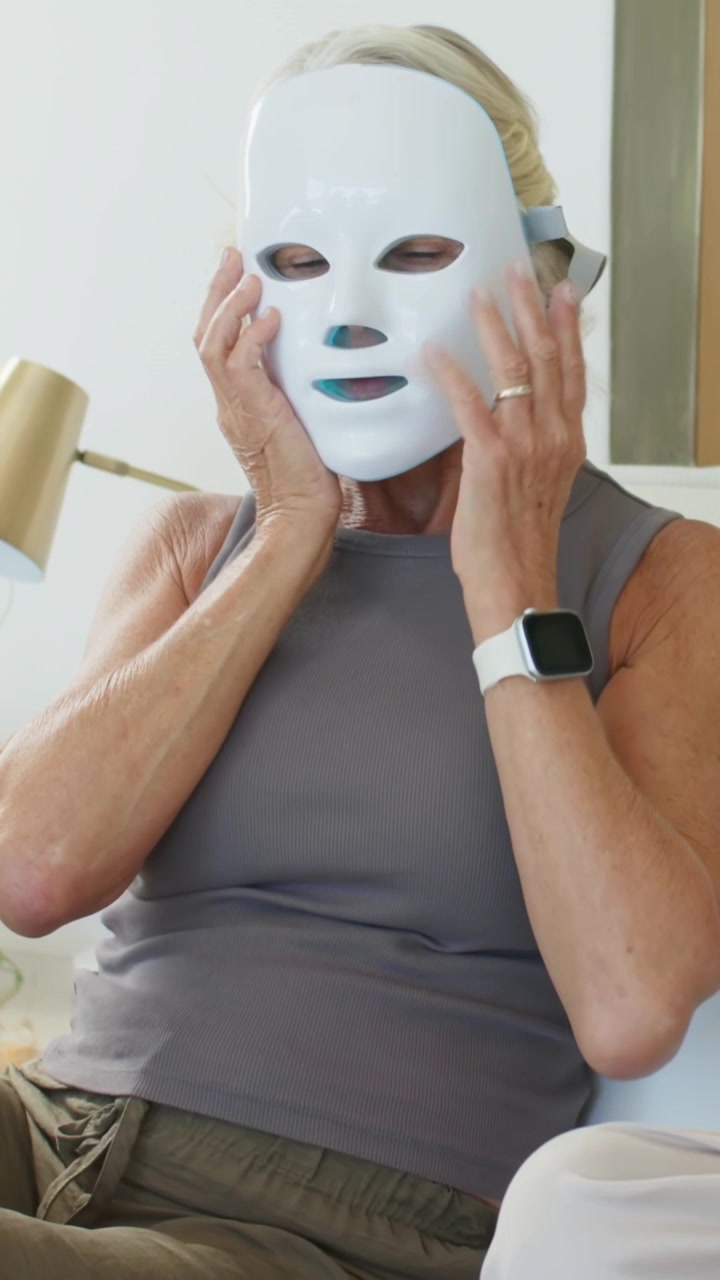Video vertical: Mujer madura sosteniendo una mascarilla LED, ajustando la correa y suavizando el ajuste para terapia de piel.