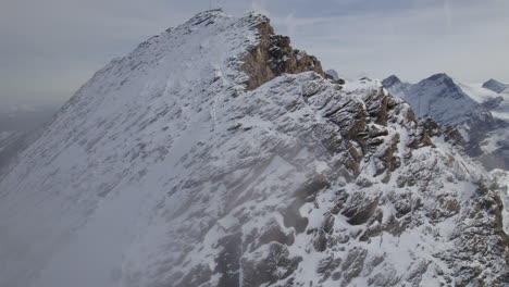 Aerial-orbit-shot-of-flight-along-rocky-and-snowy-mountain-wall-in-clouds-of-sky