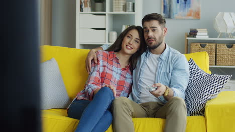 Retrato-De-Un-Joven-Y-Una-Mujer-Guapos-Viendo-La-Televisión-Juntos-Mientras-Descansan-En-El-Sofá-Amarillo-De-La-Sala-De-Estar