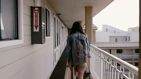 young happy tourist woman walking along american roadside hotel hall with travel bag and backpack back view slow motion.