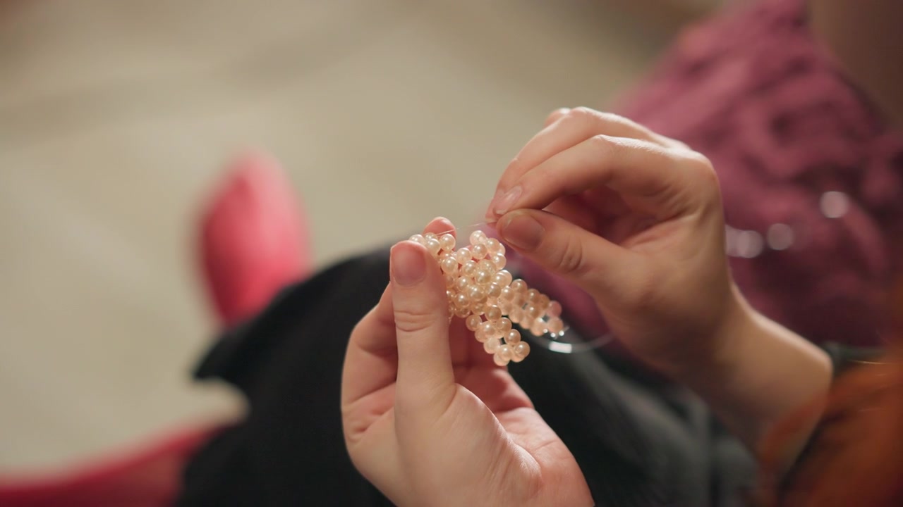 Presis smykkefremstilling. Nærbilde av hender som lager et perlearmbånd med detaljert fokus. Dyktig håndverker trer omhyggelig perler for å lage et glitrende talismanarmbånd i koselig belysning.