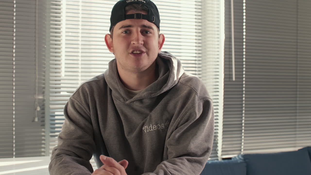 Young man in a grey hoodie sits on a stool and talks directly to the camera during an interview. Static Shot with nice even natural lighting