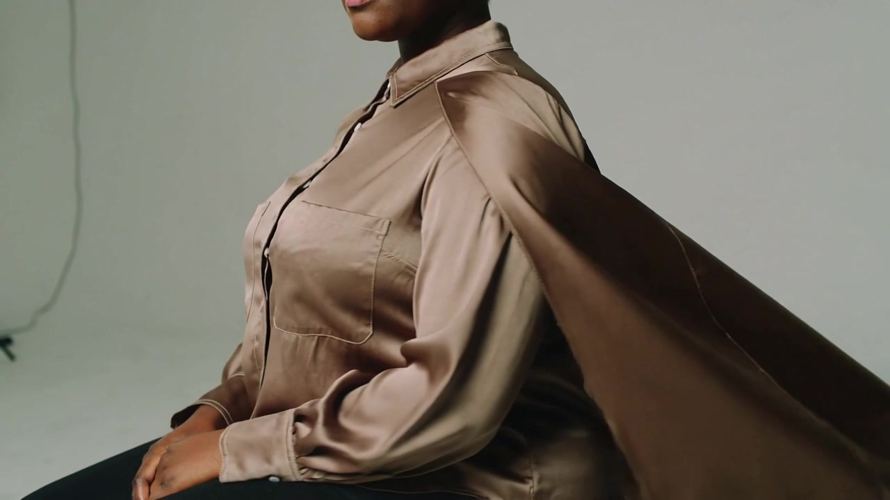 Una donna seduta in camicetta di raso che solleva un mantello di bronzo per una folata di vento di un ventilatore, girandosi di profilo in studio.