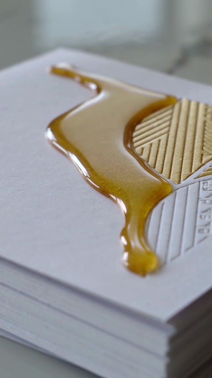 Vertical video: Falling amber liquid initiating spread over card stack, coating gold foil design