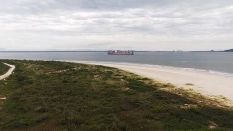 Un-Gran-Buque-De-Carga-Lleno-De-Contenedores-Navega-Frente-A-La-Costa-De-Pontal-Do-Sul-En-Paraná,-Brasil,-Drone-4k