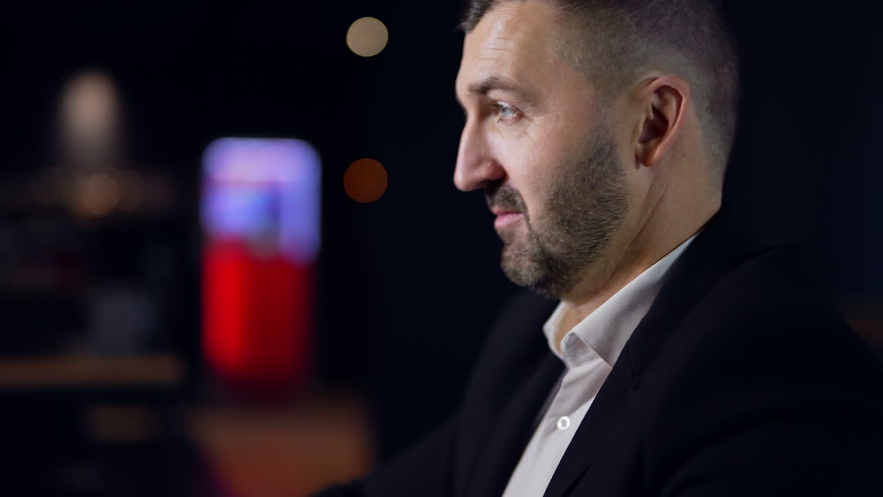 Thoughtful man in his thirties sitting in the dark room. Bearded handsome man sitting at the table quietly. Side view.