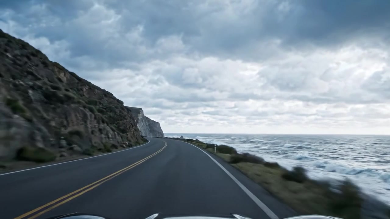 Un Sereno Viaggio Costiero: Viste accattivanti di scogliere aspre e onde che lambiscono lungo la strada tortuosa sotto cieli nuvolosi e drammatici.