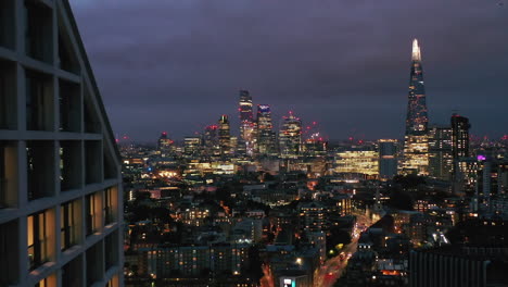 Panoramic-aerial-view-of-illuminated-downtown-skyscrapers-at-night.-Backwards-reveal-of-modern-high-rise-apartment-building.-London,-UK