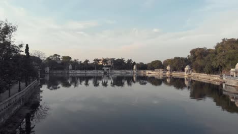 Shore-view-of-lake-Pichola-Ascending-to-Ambrai-Ghat-and-revealing-the-surrounding-Old-town,-in-Udaipur,-Rajasthan,-India---Aerial-ascending-Fly-over-reveal-shot