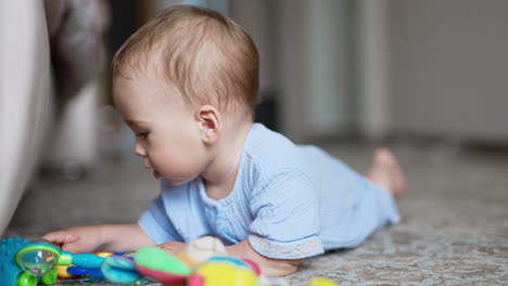nice kid wearing blue shirt lies on the floor. little boy touches toys lying beside him. close up. blurred backdrop.