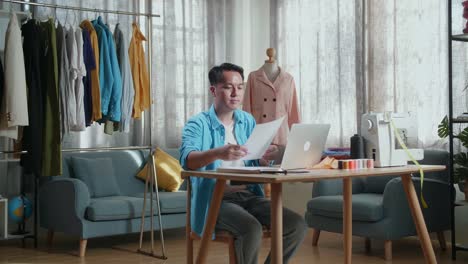 asian male designer with sewing machine designing clothes on paper while working with laptop computer in the studio