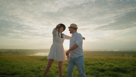 los amantes sonrientes bailan alegremente en una colina cubierta de hierba al atardecer cerca de un lago tranquilo, moviendo juguetonamente sus manos. el hombre con una camisa blanca y un sombrero y la mujer con un vestido blanco abrazan la luz dorada