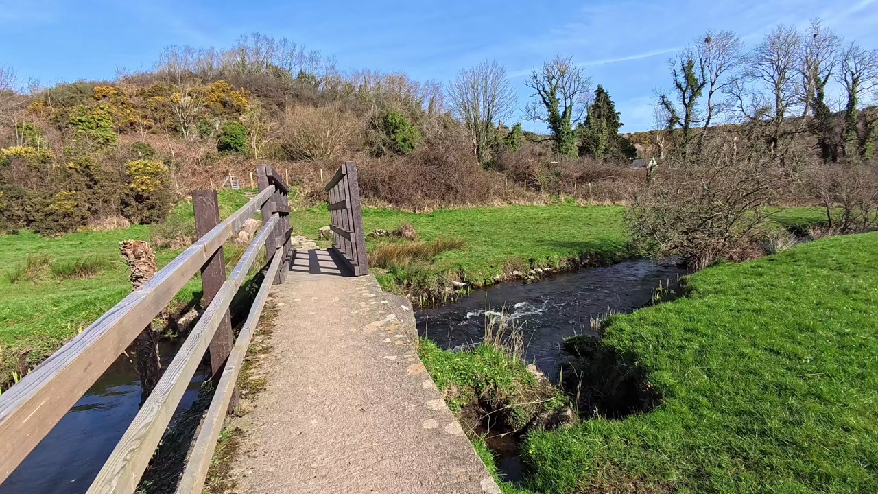 Walking towards sunlit wooden bridge crossing over flowing clean stream in welsh countryside meadow