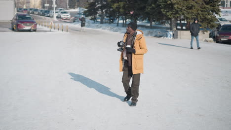 journalist walking along snowy city street holding coffee cup and camera, glancing upward, lined with pine trees, as parked and passing cars create urban winter scene under soft daylight shadows