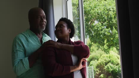 Afroamerikanisches-Seniorenpaar-Umarmt-Sich,-Während-Es-Zu-Hause-Aus-Dem-Fenster-Schaut