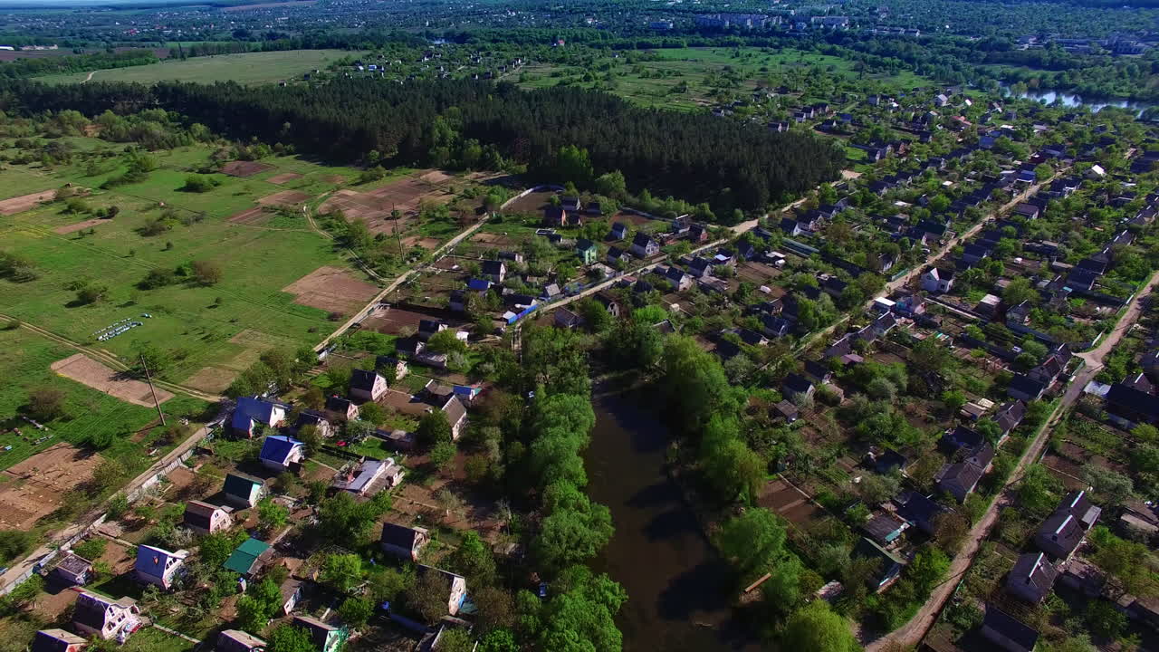 Panorama of the beautiful green village on the river bank. Flight over the shining water on sunny day. Big city at backdrop.