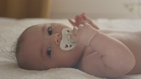 Baby-with-playful-pacifier,-making-eye-contact-with-camera-in-white-sunlit-bedroom