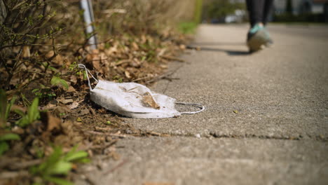 a discarded, dirty face mask left on the sidewalk as a person walks towards camera and past it