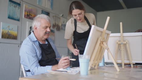 a female teacher shows a retired man how to draw a picture with paints and a brush at courses for the elderly. a senior man draws a picture to a group of pensioners