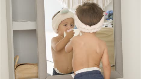 little latin baby boy shirtless with blue shorts and wearing a funny hair band with ears playing with makeup sponges looking at himself on the mirror