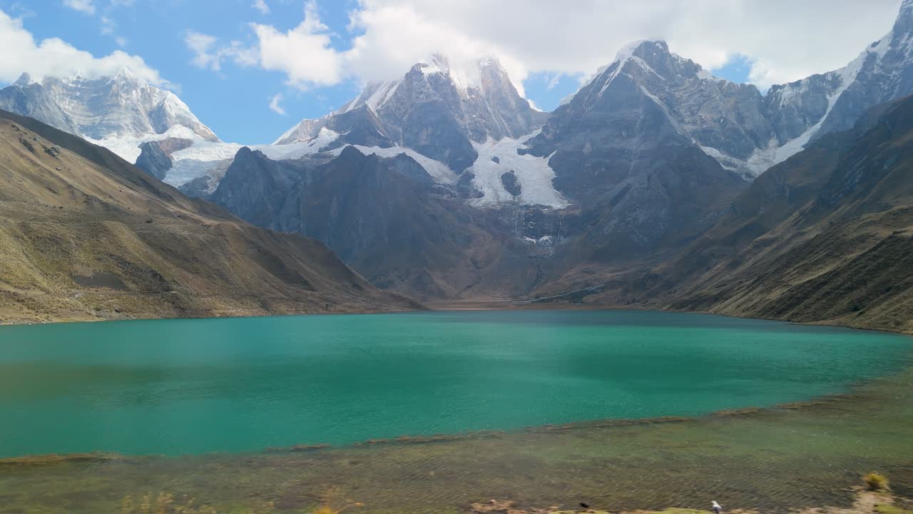 A breathtaking aerial trucking shot glides along a vivid turquoise glacial lagoon, framed by towering peaks of Peru’s stunning Cordillera Huayhuash trek