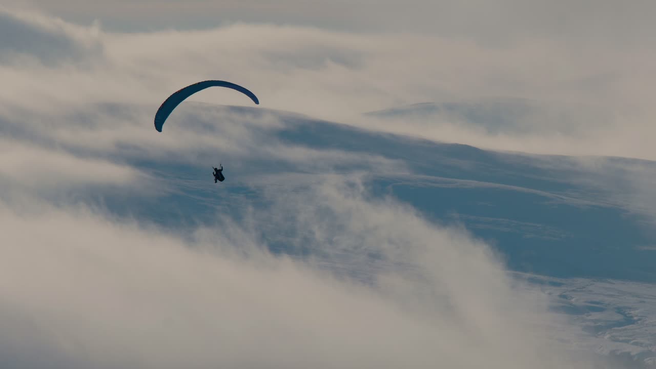 Paraglider shot against the light with snow covered hills and clouds in the background, Cumbria UK