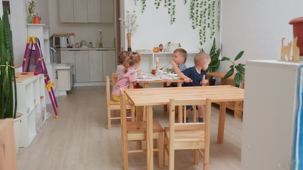 Anak-anak prasekolah duduk bersama di ruang kelas taman kanak-kanak yang nyaman, menikmati makanan sementara seorang wanita menghampiri untuk memeriksa mereka di bawah cahaya siang yang hangat dikelilingi oleh furnitur kayu