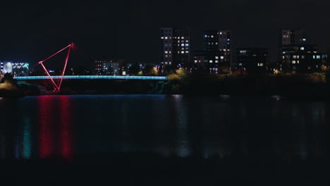 Night-view-of-Pae-park-pedestrian-bridge-illuminated-with-colorful-light-over-lake-water-with-buildings-at-background,-Tallinn,-Estonia