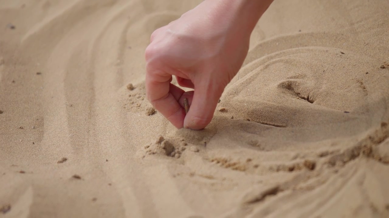 Närbild av en hand som använder ett litet föremål för att rita cirkulära former i strandsand, vilket fångar en enkel, kreativ och avkopplande utomhusaktivitet