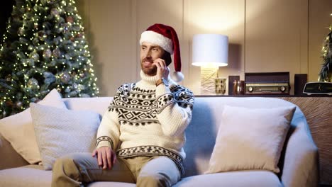 un hombre alegre y guapo con sombrero de santa sentado en casa con un árbol de navidad y charlando en un smartphone felicitando a sus amigos con las vacaciones