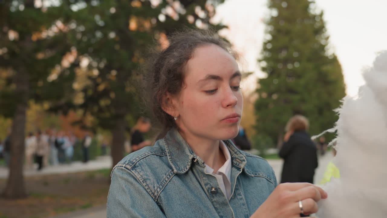 Donna bianca che addenta zucchero filato con gioia, folla vivace nel parco, giacca di jeans, bocca che ride, mani appiccicose, bagliore del tramonto, bancarella di un venditore nelle vicinanze, energia giocosa, scena di celebrazione spontanea che evoca la famiglia.