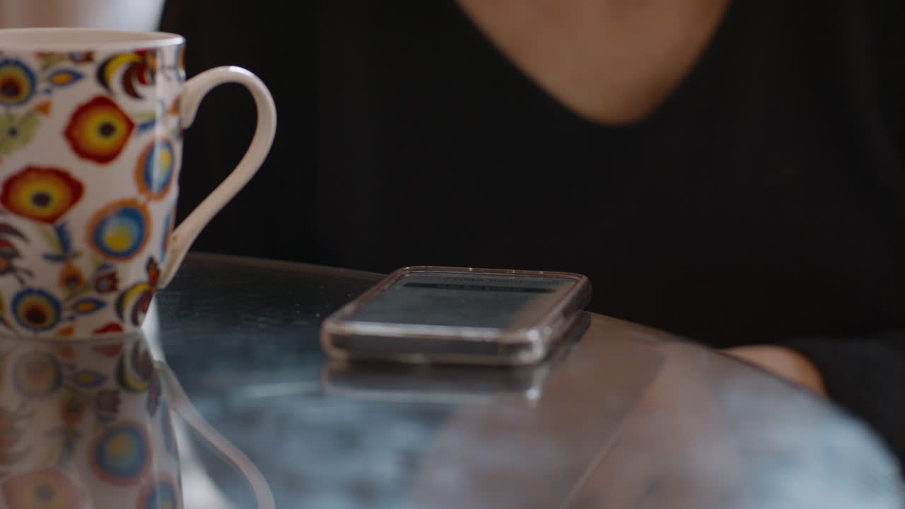 A close-up shot of a smartphone resting face-up on a shiny glass top kitchen table beside a vibrant, patterned coffee mug. A woman in a black V-neck top sits nearby, her hand subtly blurred in motion.