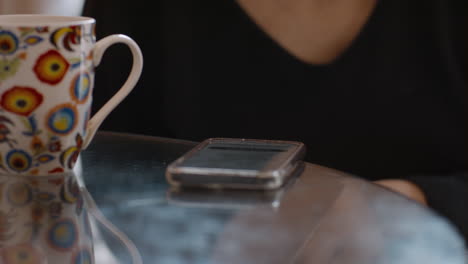 a close-up shot of a smartphone resting face-up on a shiny glass top kitchen table beside a vibrant, patterned coffee mug. a woman in a black v-neck top sits nearby, her hand subtly blurred in motion.