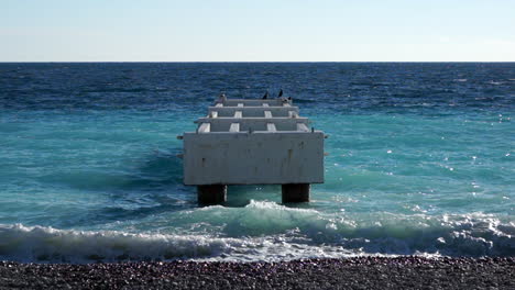 Symmetric-shot-of-an-old-jetty-in-a-blue-sea,-seagulls-flying-by-SLOMO